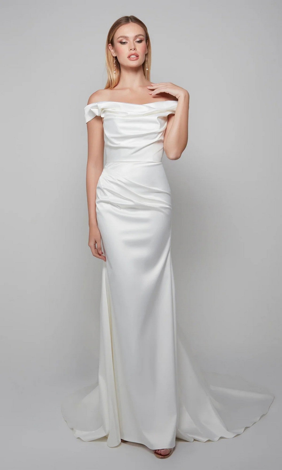 A woman stands against a plain light gray background wearing the Long White Dress: Alyce 7064, an ivory, floor-length off-the-shoulder gown.
